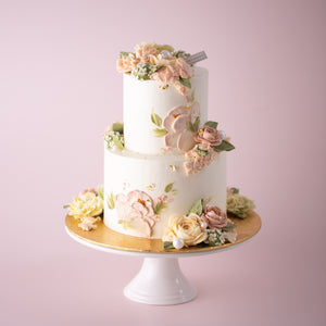 
                  
                    Load image into Gallery viewer, A two tier cake with a white buttercream base. The cake has both hand piped buttercream flowers and palette knife buttercream flowers. The flowers are in shades if ivory and pastel pink, and look incredibly realistic.
                  
                