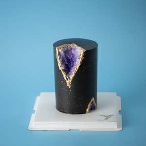 
                  
                    Load image into Gallery viewer, A tall and slim cake, this cake is covered in a dark bluish purple fondant. The fondant has speckles of gold splattered on it that resemble stars. A chunk of the cake has been cut out adn replaced with edible purple rock candy that resemble Amethyst gems, so the cake looks like a geode.
                  
                