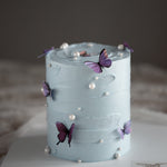 A cake with light blue buttercream scraped to have a swirl-like texture. The cake has multiple edible wafer butterflies in purple and pink placed around the cake, and it also has white edible sugar pearls scattered around.