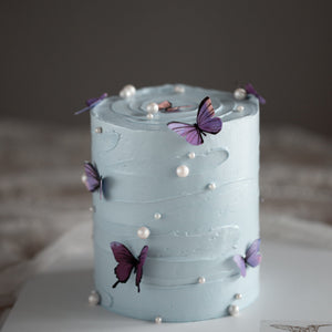 
                  
                    Load image into Gallery viewer, A cake with light blue buttercream scraped to have a swirl-like texture. The cake has multiple edible wafer butterflies in purple and pink placed around the cake, and it also has white edible sugar pearls scattered around.
                  
                