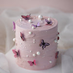 A cake with pink buttercream scraped to have a swirl-like texture. The cake has multiple edible wafer butterflies in purple and pink placed around the cake, and it also has white edible sugar pearls scattered around.