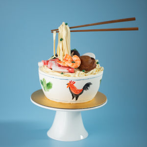 
                  
                    Load image into Gallery viewer, A cake that has been decorated to look like a bowl of noodles. There are fondant shrimp, sliced meat, abalone and spring onions on top of the noodles. Floating above in the :anti gravity&amp;quot; style is a pair of chopsticks also made of fondant, holding into the fondant noodles.
                  
                