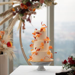 A three tier wedding cake with a white buttercream base. The cake has red, orange and yellow edible sugar leaves that spiral around and upwards the cake, resembling leaves blowing in an autumn breeze. 