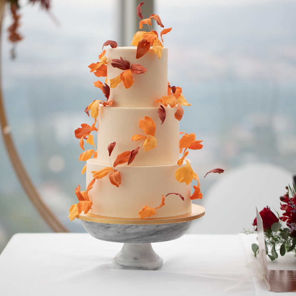 
                  
                    Load image into Gallery viewer, A three tier wedding cake with a white buttercream base. The cake has red, orange and yellow edible sugar leaves that spiral around and upwards the cake, resembling leaves blowing in an autumn breeze.
                  
                