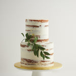 A two tier naked wedding cake, with white buttercream as a rough outer coating. This rustic wedding cake has a thick sprig of edible green sugar leaves gently hanging from the side. The cake also has bits of gold leaf scattered around the cake delicately.