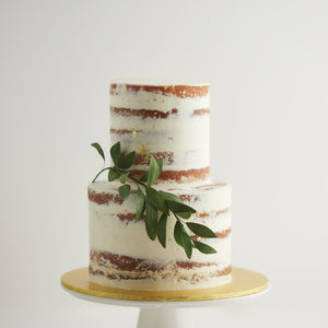 
                  
                    Load image into Gallery viewer, A two tier naked wedding cake, with white buttercream as a rough outer coating. This rustic wedding cake has a thick sprig of edible green sugar leaves gently hanging from the side. The cake also has bits of gold leaf scattered around the cake delicately.
                  
                