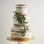 A three tier naked wedding cake, with white buttercream as a rough outer coating. This rustic wedding cake has a thick sprig of edible green sugar leaves gently hanging from the side. The cake also has bits of gold leaf scattered around the cake delicately.