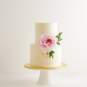 
                  
                    Load image into Gallery viewer, A two tier wedding cake with a white buttercream base. There is one large light pink edible sugar peonies on the cake, along with some green edible sugar leaves. The flowers look incredibly realistic. The cake also has some gold leaf that has been carefully applied on it.
                  
                