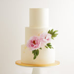 A three tier wedding cake with a white buttercream base. There are two large light pink edible sugar peonies on the cake, along with some green edible sugar leaves. The flowers look incredibly realistic. The cake also has some gold leaf that has been carefully applied on it.