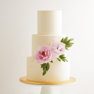 
                  
                    Load image into Gallery viewer, A three tier wedding cake with a white buttercream base. There are two large light pink edible sugar peonies on the cake, along with some green edible sugar leaves. The flowers look incredibly realistic. The cake also has some gold leaf that has been carefully applied on it.
                  
                