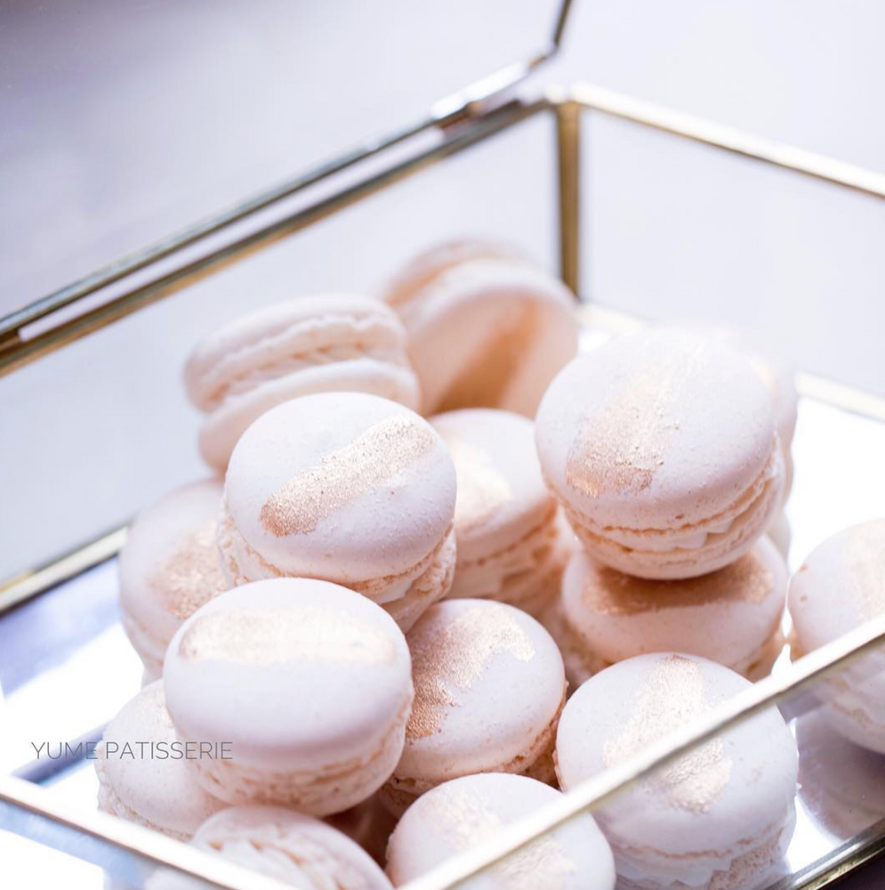 A transparent box filled with small light pink macarons. The macarons have a single streak of gold painted on their top shells, giving it an elegant, classy look