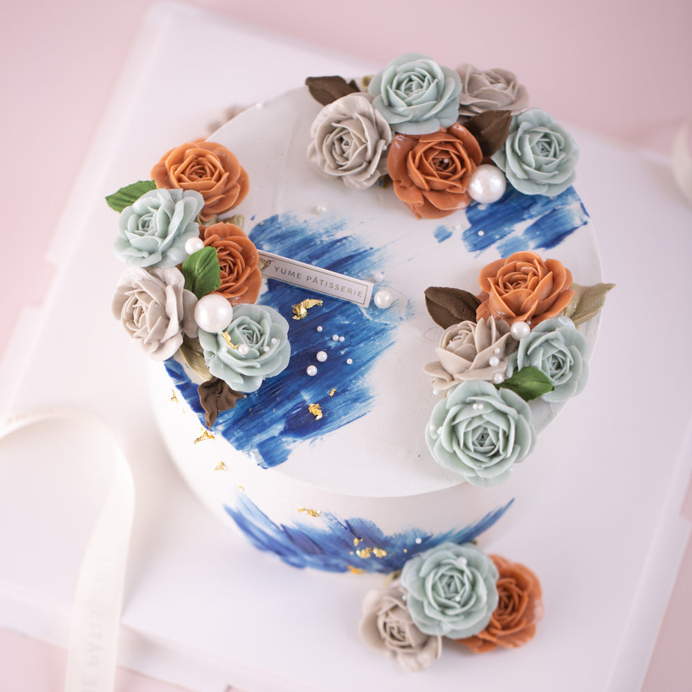 A white buttercream cake with accents of blue buttercream. The cake has muted orange, purple and blue buttercream flowers, and bits of gold leaf.