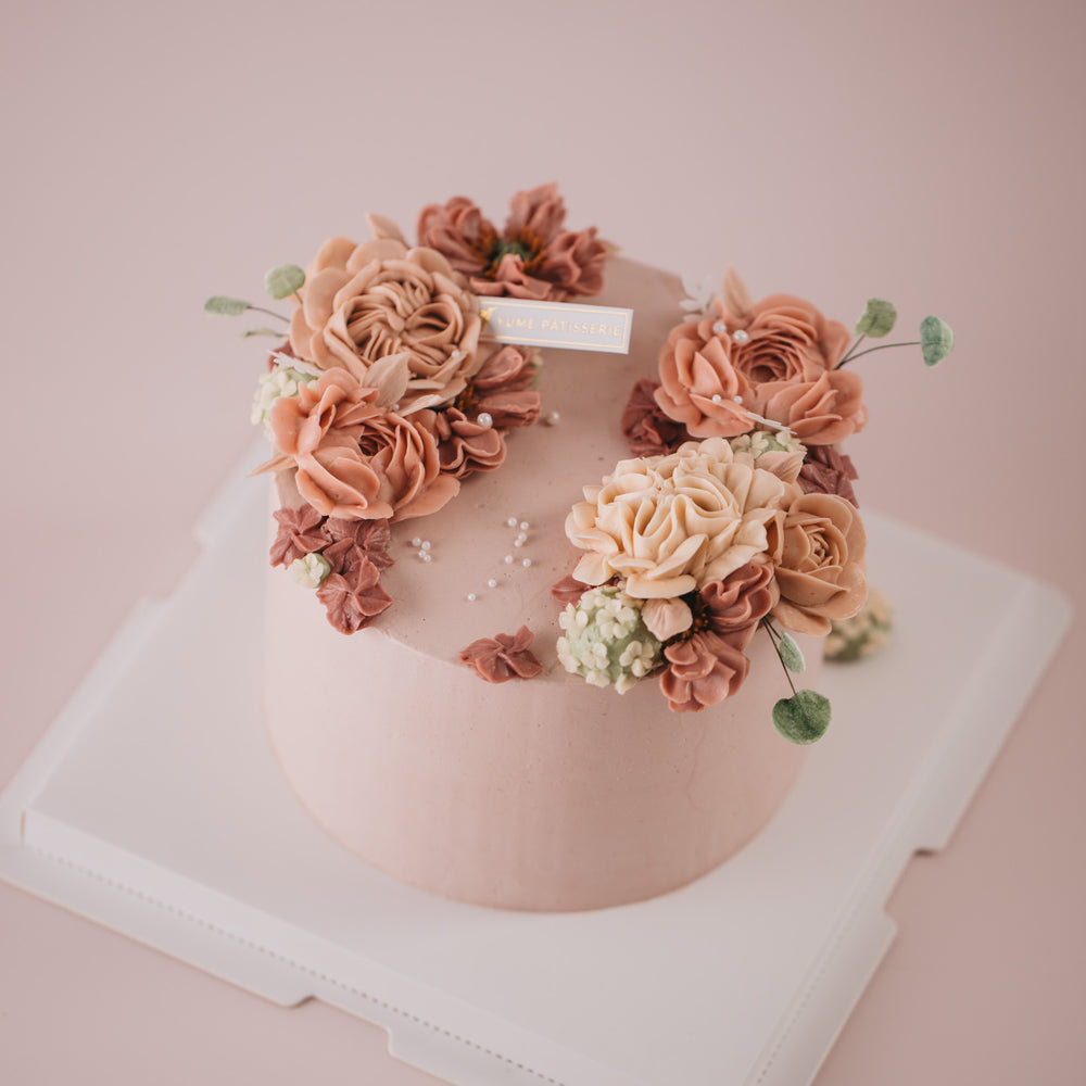 A light pink. buttercream base, with pink, beige and ivory buttercream flowers on top in a wreath style.
