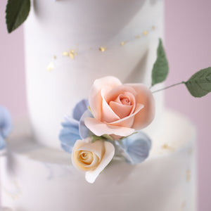 
                  
                    Load image into Gallery viewer, A close up on the flowers on the middle of the cake, between the two tiers. There are two edible sugar roses, one light pink and one ivory. There are also wafer hydrangeas in blue for that colour contrast behind the roses.
                  
                