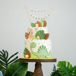 Two tier dinosaur themed cake. This cake has a white and green buttercream base, and many little tropical ferns in shades of green and brown for the Jurassic Park effect. There are two fondant dinosaurs on the cake, and many little fondant dino eggs around.