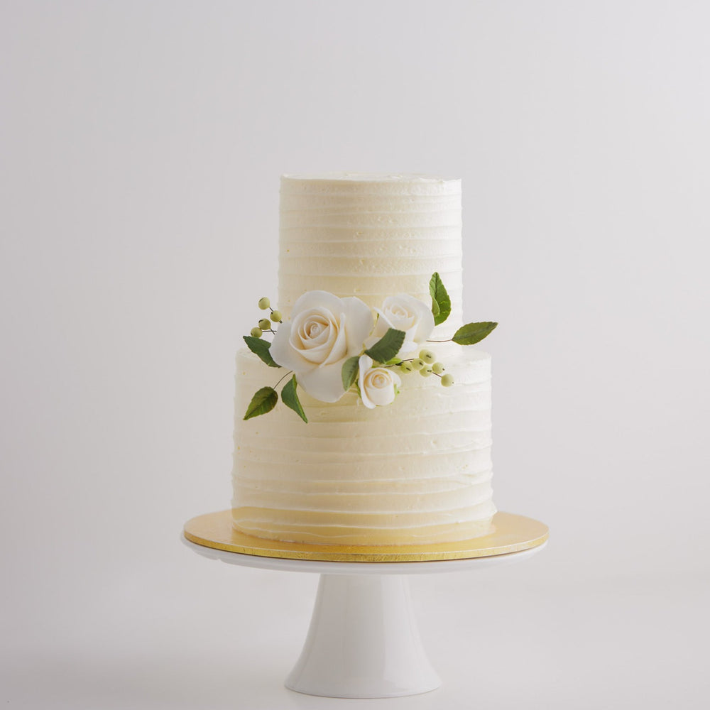  A two tier white buttercream cake with textured lines that go across horizontally. The middle tier has some white edible sugar roses, with deep green sugar leaves for a pop of colour. The roses are incredibly realistic.