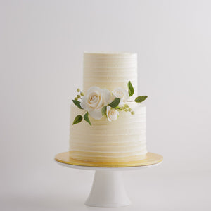 
                  
                    Load image into Gallery viewer,  A two tier white buttercream cake with textured lines that go across horizontally. The middle tier has some white edible sugar roses, with deep green sugar leaves for a pop of colour. The roses are incredibly realistic.
                  
                