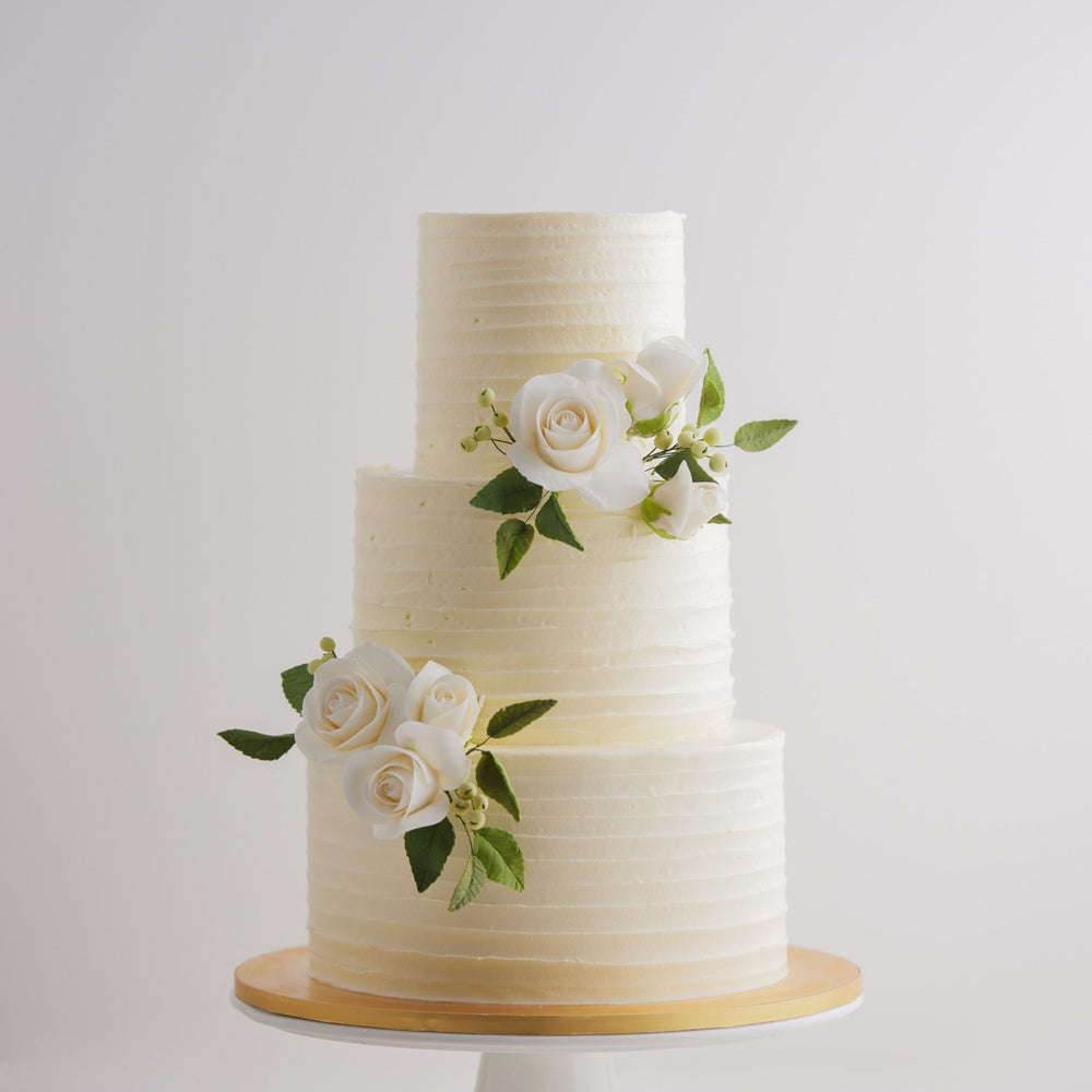 A three tier white buttercream cake with textured lines that go across horizontally. the top and bottom of the middle tier have some white edible sugar roses, with deep green sugar leaves for a pop of colour. The roses are incredibly realistic. 