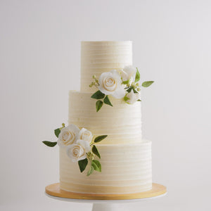 
                  
                    Load image into Gallery viewer, A three tier white buttercream cake with textured lines that go across horizontally. the top and bottom of the middle tier have some white edible sugar roses, with deep green sugar leaves for a pop of colour. The roses are incredibly realistic. 
                  
                