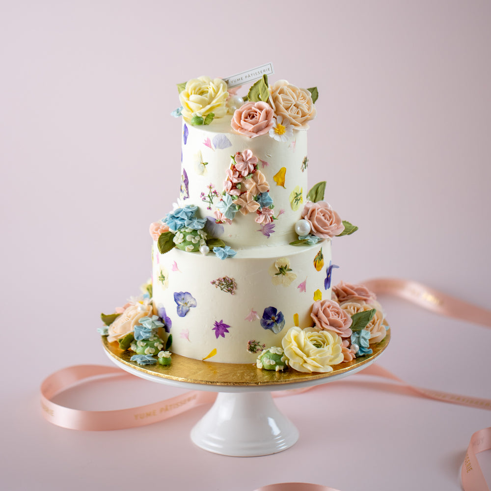 A two tier cake with colourful edible flowers pressed into the buttercream. The cake has a white buttercream base. The top tier has pastel buttercream flowers forming the letter 
