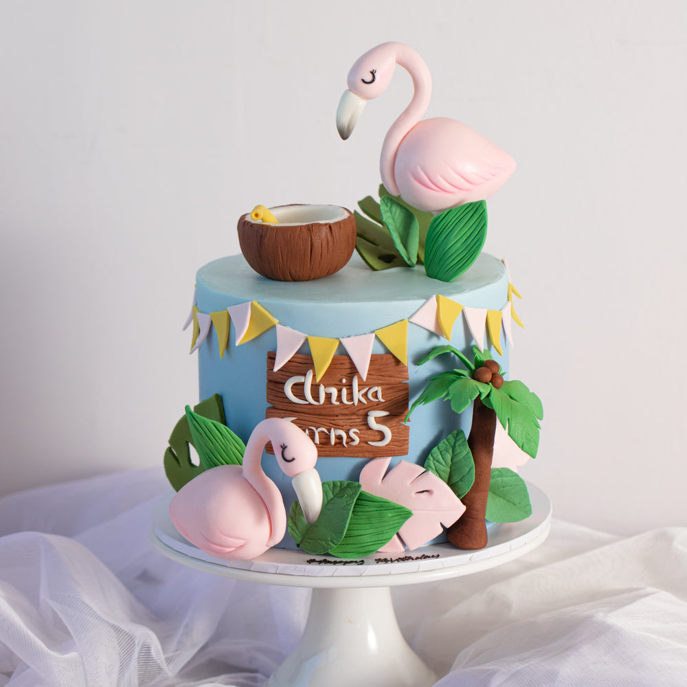 A tropical themed cake decorated with two fondant flamingos. The top of the cake has yellow and light pink bunting made of fondant. The base of the cake is a light blue. There are many 3D fondant decorations around the cake to give it a lovely, full look.