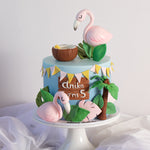 A tropical themed cake decorated with two fondant flamingos. The top of the cake has yellow and light pink bunting made of fondant. The base of the cake is a light blue. There are many 3D fondant decorations around the cake to give it a lovely, full look.