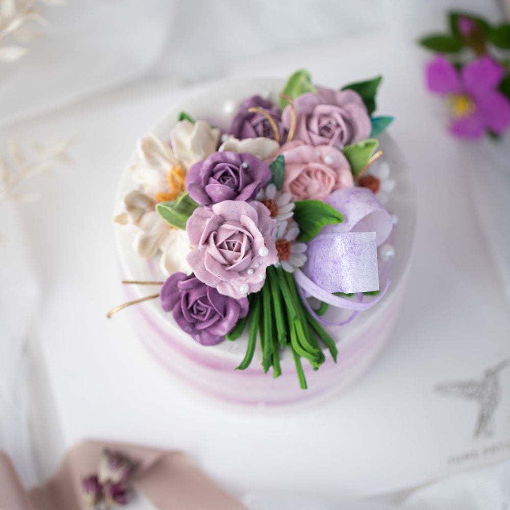 
                  
                    Load image into Gallery viewer, A white buttercream cake with streaks of purple buttercream, and various shades of purple buttercream roses on top of the cake. The buttercream roses have green stems, and resemble a bouquet of flowers. The cake has some edible sugar pearls scattered on top.
                  
                