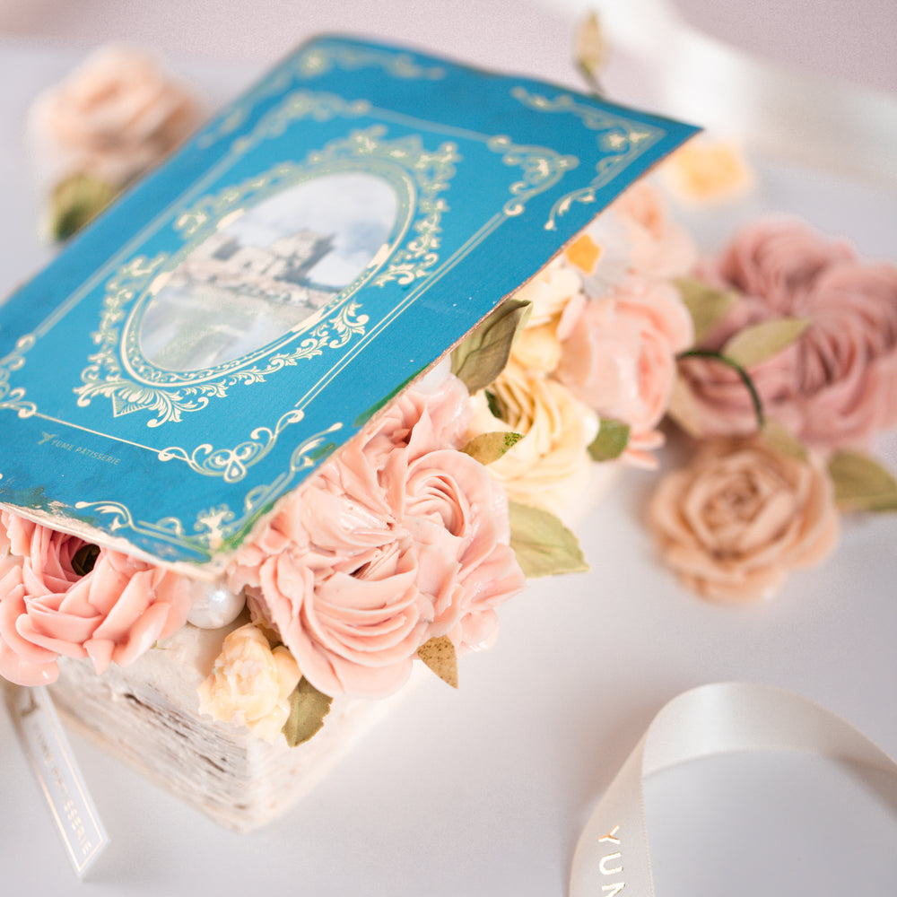 
                  
                    Load image into Gallery viewer, A cake that has been decorated to look like q vintage fairytale book. The book cover is partially open, and there are buttercream flowers spilling out. The boom cover is blue, and the buttercream flowers are shades of coral and ivory
                  
                