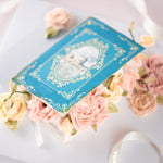 A cake that has been decorated to look like q vintage fairytale book. The book cover is partially open, and there are buttercream flowers spilling out. The boom cover is blue, and the buttercream flowers are shades of coral and ivory