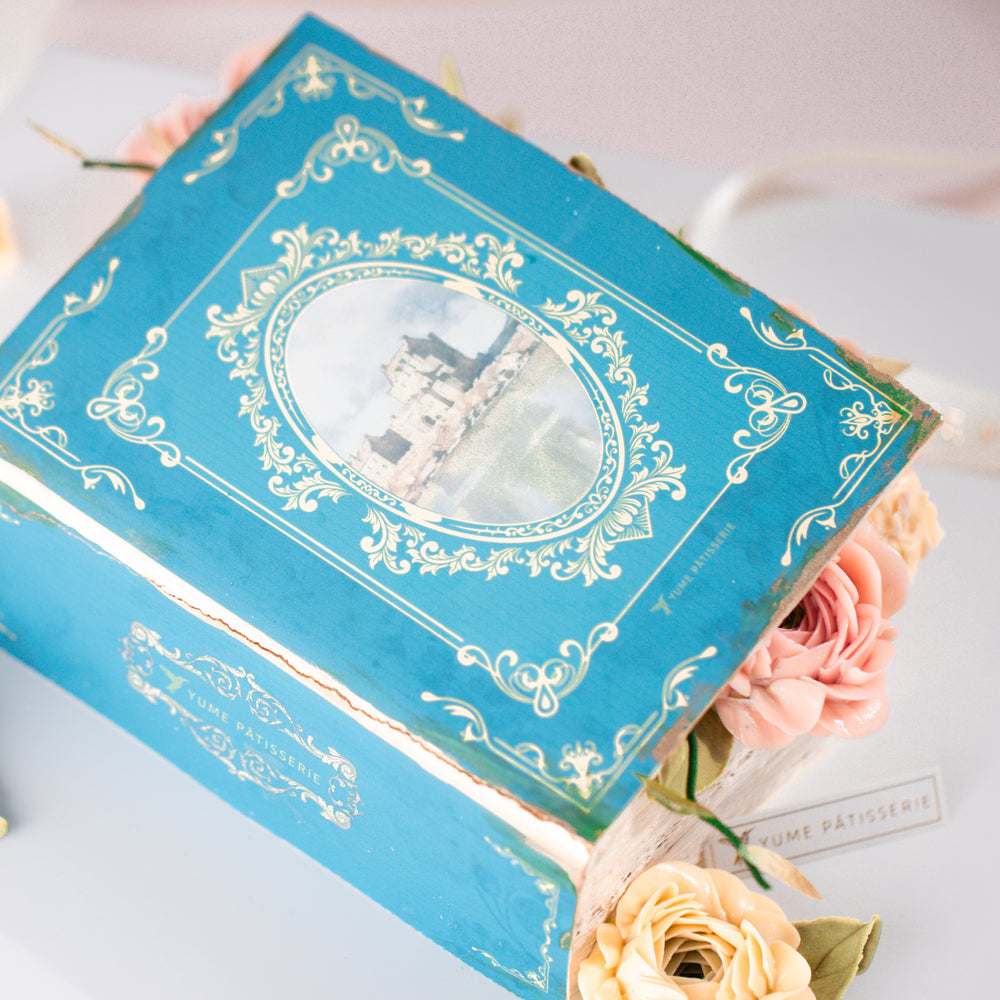 
                  
                    Load image into Gallery viewer, A cake that has been decorated to look like q vintage fairytale book. The book cover is partially open, and there are buttercream flowers spilling out. The boom cover is blue, and the buttercream flowers are shades of coral and ivory
                  
                