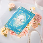 A cake that has been decorated to look like q vintage fairytale book. The book cover is partially open, and there are buttercream flowers spilling out. The boom cover is blue, and the buttercream flowers are shades of coral and ivory.