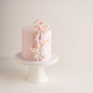 
                  
                    Load image into Gallery viewer, A light ponk fondant base on a tall cake, with delicate white edible wafer paper scallops cascading down one side of the cake. There are small edible wafer sakura flowers on the cake, giving it a Japanese vibe.
                  
                