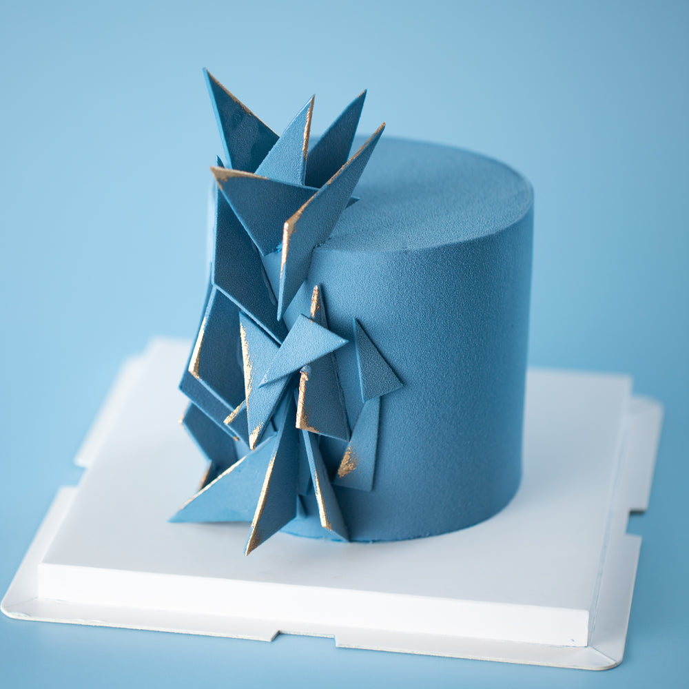 A cake that has been airbrushed with dark blue velour chocolate spray. The cake has many blue fondant triangles sticking out from the side, like shards of glass. The edges of the fondant triangles have been painted gold for a contrasting accent.