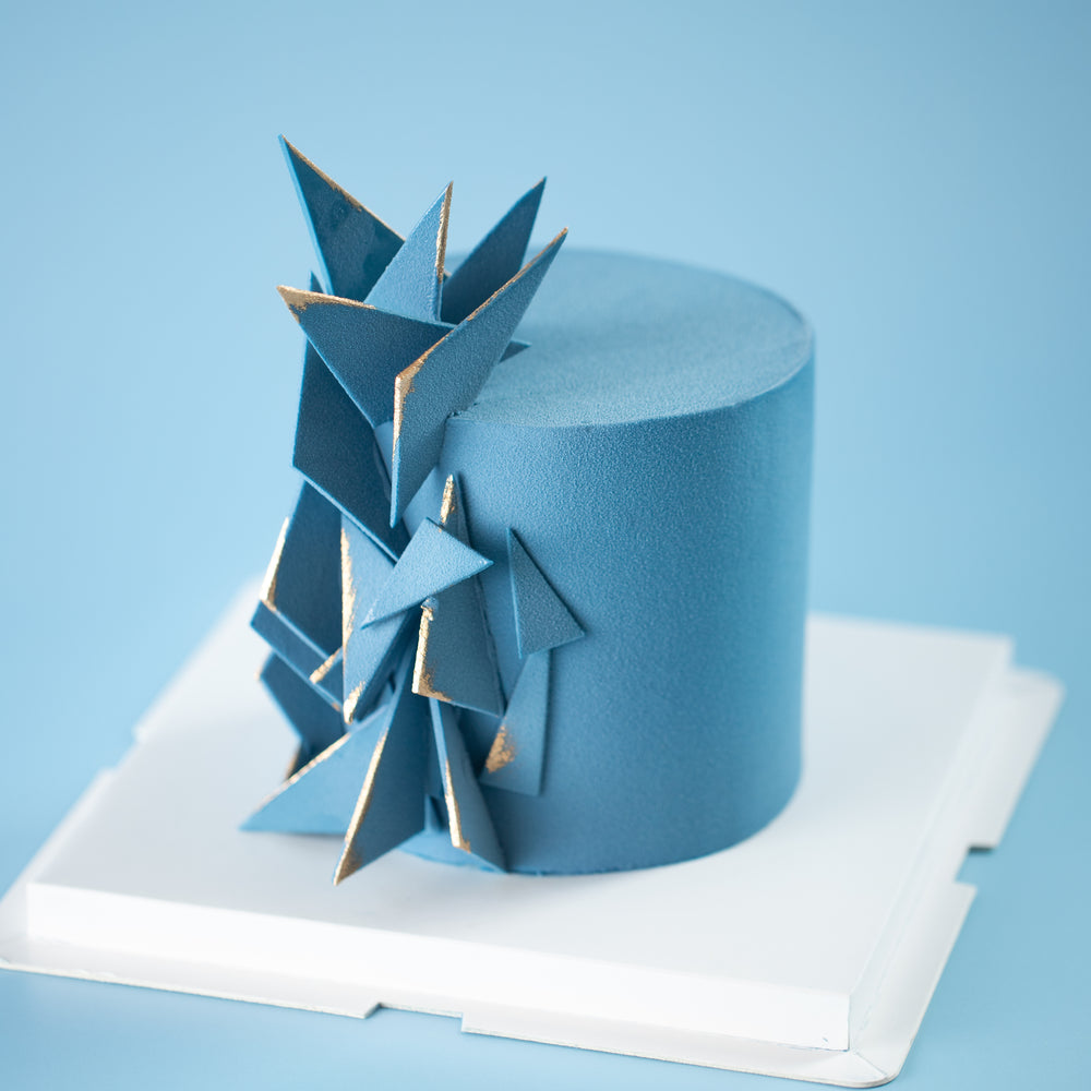 A cake that has been airbrushed with dark blue velour chocolate spray. The cake has many blue fondant triangles sticking out from the side, like shards of glass. The edges of the fondant triangles have been painted gold for a contrasting accent.