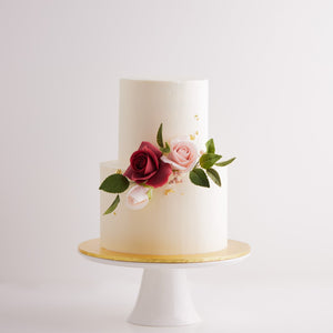 
                  
                    Load image into Gallery viewer, A two tier wedding cake with a smooth white buttercream base. The cake has pink and red edible sugar roses on it, along with some green edible sugar leaves. The roses look incredibly realistic. The white buttercream also has some flakes of gold leaf carefully placed on them.
                  
                