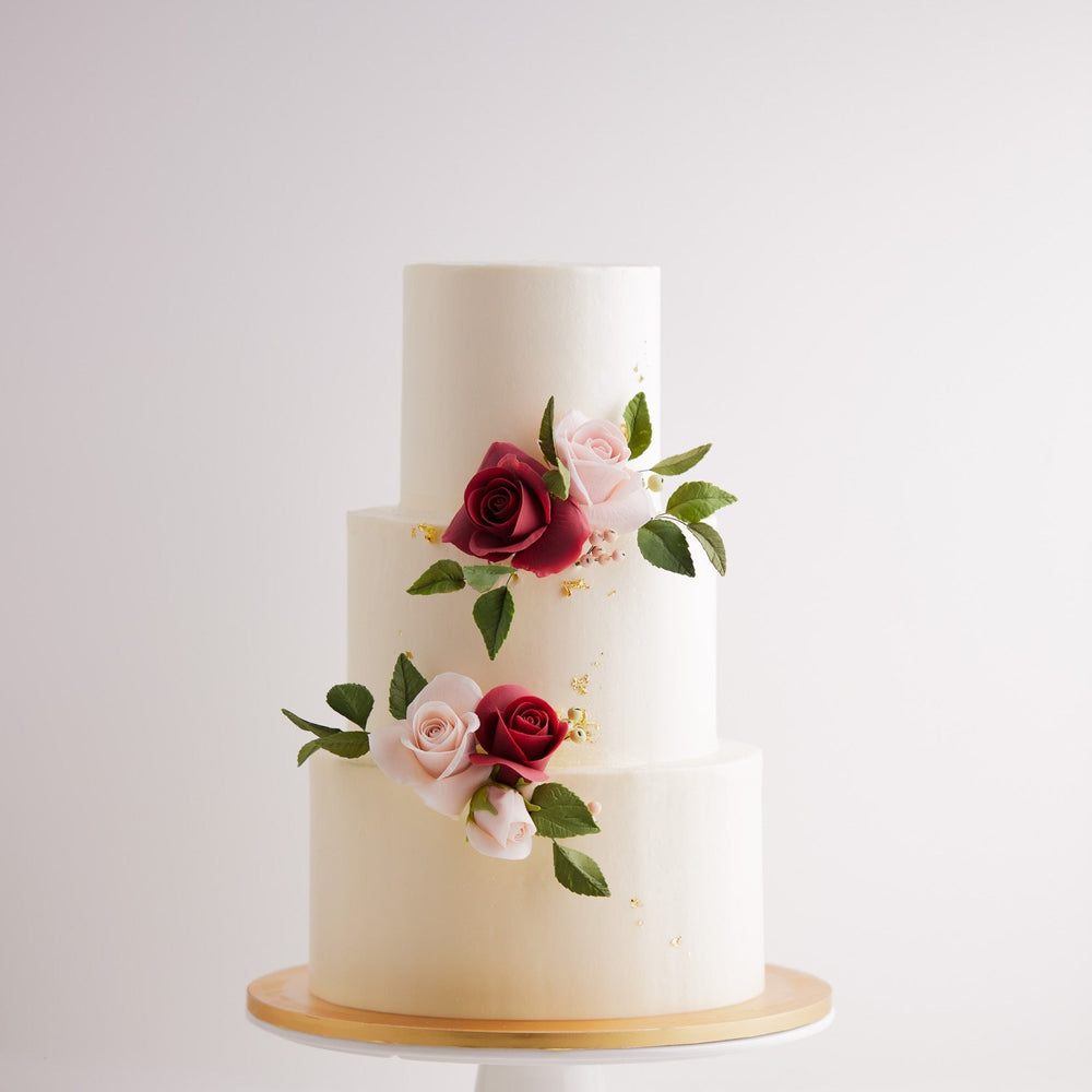 A three tier wedding cake with a smooth white buttercream base. The cake has pink and red edible sugar roses on it, along with some green edible sugar leaves. The roses look incredibly realistic. The white buttercream also has some flakes of gold leaf carefully placed on them.