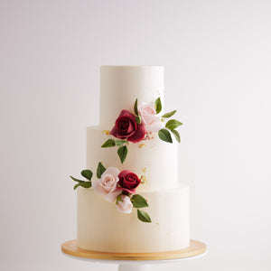
                  
                    Load image into Gallery viewer, A three tier wedding cake with a smooth white buttercream base. The cake has pink and red edible sugar roses on it, along with some green edible sugar leaves. The roses look incredibly realistic. The white buttercream also has some flakes of gold leaf carefully placed on them.
                  
                