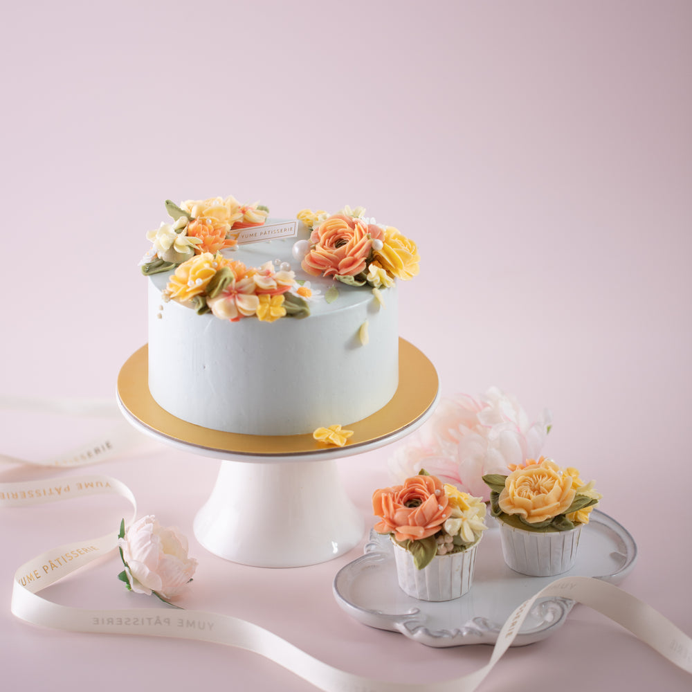 A pale blue cake with bright coral and yellow hand piped buttercream flowers. The cake also has some sugar pearls delicately scattered on top. The flowers look incredibly lifelike. Next to the cake, there are some matching cupcakes with the same yellow and coral buttercream flowers.