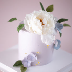 
                  
                    Load image into Gallery viewer, A cake with a lilac buttercream base. The top of the cake has a large white wafer peony on it, along with some dark green edible sugar leaves. On the sides of the cake, there are a couple of bluish purple hydrangea petals, and some gold leaf. All the flowers look incredibly realistic.
                  
                