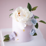 A cake with a lilac buttercream base. The top of the cake has a large white wafer peony on it, along with some dark green edible sugar leaves. On the sides of the cake, there are a couple of bluish purple hydrangea petals, and some gold leaf. All the flowers look incredibly realistic.