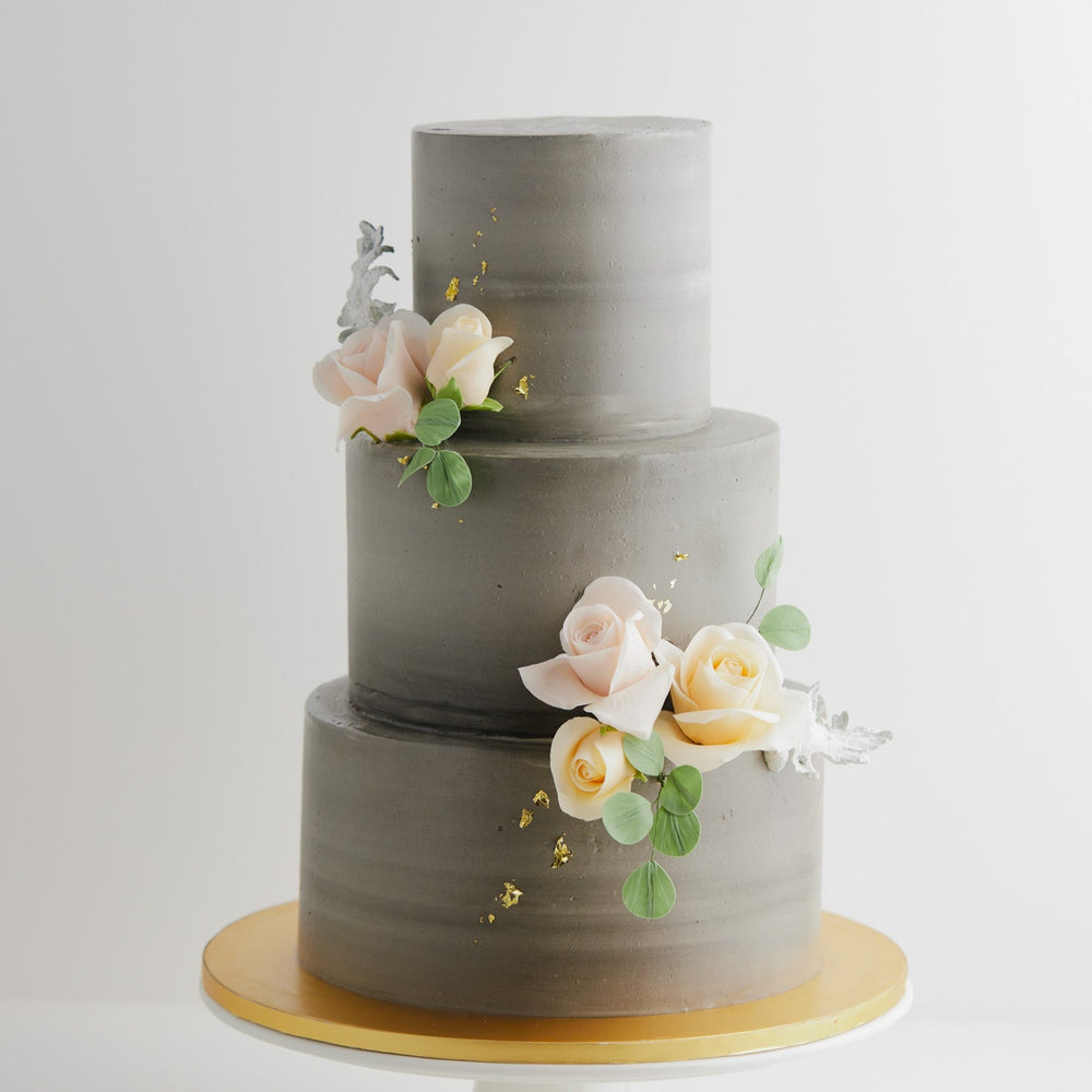 A three tier wedding cake with a concrete grey buttercream base. The buttercream has streaks of lighter grey marbled into it. There are some light pink and ivory edible sugar roses in the middle, along with some pale edible green sugar leaves. The flowers look incredibly realistic.