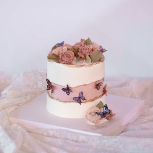 
                  
                    Load image into Gallery viewer, A white buttercream faultline cake, where the &amp;quot;inner&amp;quot; part is a light shade of blush. There are pink and purple edible wafer butterflies in the middle, and the edges of the faultline are painted with gold luster dust. The top of the cake is covered in many hand piped pink buttercream roses.
                  
                