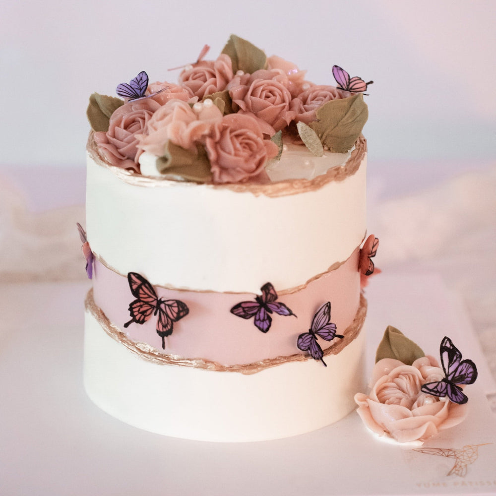 
                  
                    Load image into Gallery viewer, A white buttercream faultline cake, where the &amp;quot;inner&amp;quot; part is a light shade of blush. There are pink and purple edible wafer butterflies in the middle, and the edges of the faultline are painted with gold luster dust. The top of the cake is covered in many hand piped pink buttercream roses.
                  
                