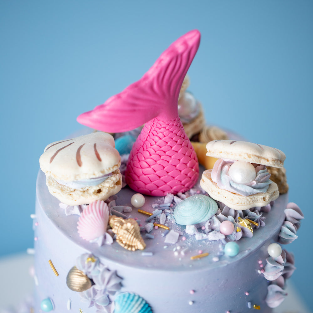 A light purple and blue buttercream cake with numerous sprinkles carefully scattered around the cake. The cake has several ivory macaron seashells, and also has a single bright pink chocolate mermaid tail sticking out from the middle. There are many tiny fondant seashells in gold, blue and purple placed around the top and bottom of the cake.