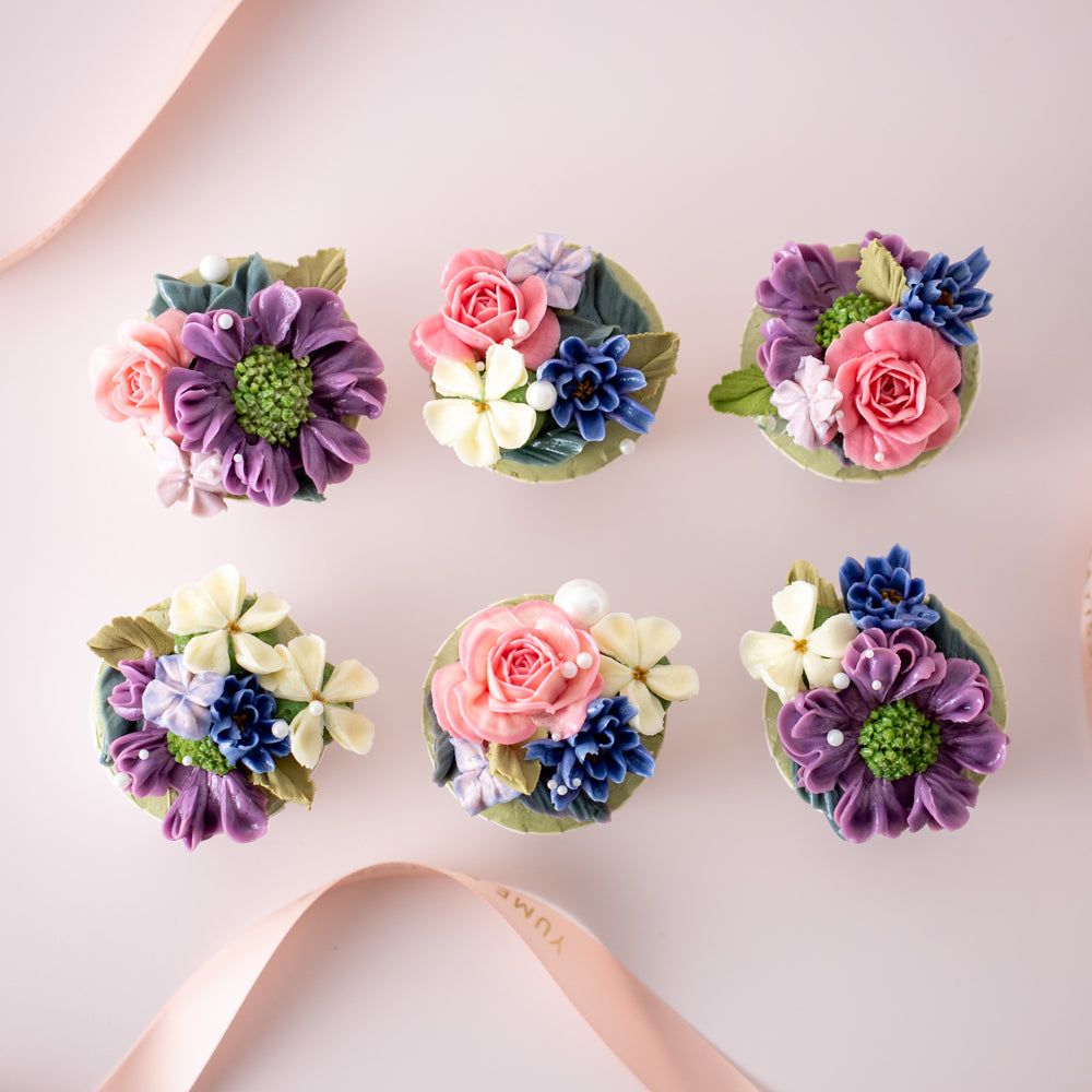6 cupcakes with assorted hand piped buttercream flowers in vibrant pink, purple, blue and ivory. There are sugar pearls lightly sprinkled on top. The flowers look incredibly realistic and beautiful.