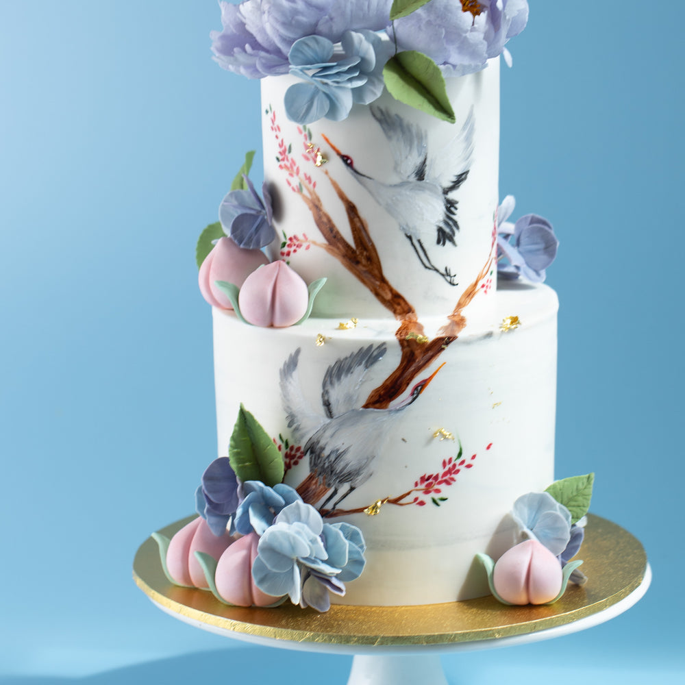 
                  
                    Load image into Gallery viewer, A close up on the bottom tier of the cake. There are pink fondant peaches around the base of the cake, and  a handpainted crane in the middle flying upwards.
                  
                