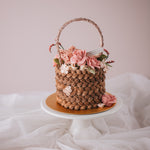 A cake that has been decorated to look like a flower basket. The outside has light brown buttercream that has been piped to resemble a woven basket, and it has assorted buttercream flowers in pink, ivory and white spilling out of it. It is a realistic flower basket cake.