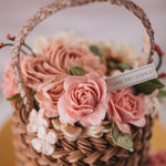A cake that has been decorated to look like a flower basket. The outside has light brown buttercream that has been piped to resemble a woven basket, and it has assorted buttercream flowers in pink, ivory and white spilling out of it. It is a realistic flower basket cake.