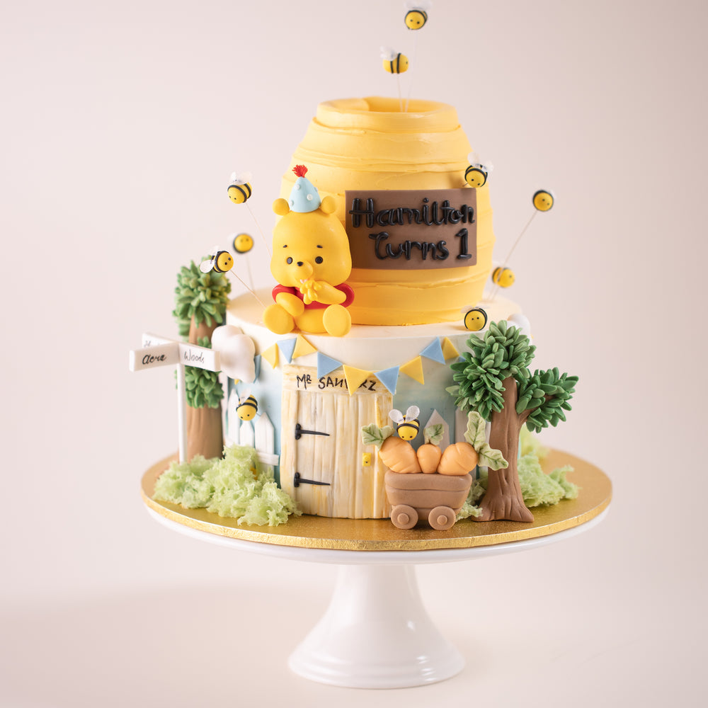 A two tier Winnie the Pooh inspired cake. The top of the cake is carved into the shape of a honey pot.