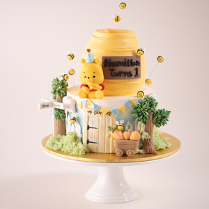 
                  
                    Load image into Gallery viewer, A two tier Winnie the Pooh inspired cake. The top of the cake is carved into the shape of a honey pot.
                  
                